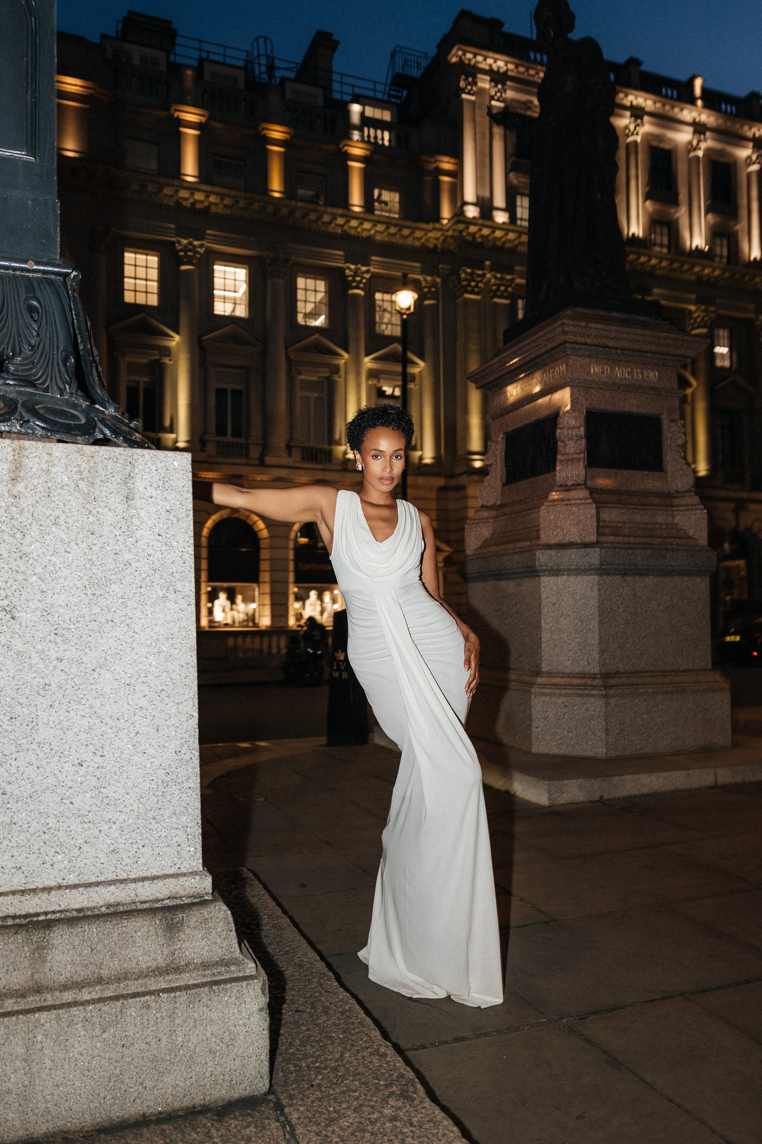 Woman in a white dress standing in an urban setting with illuminated buildings.
