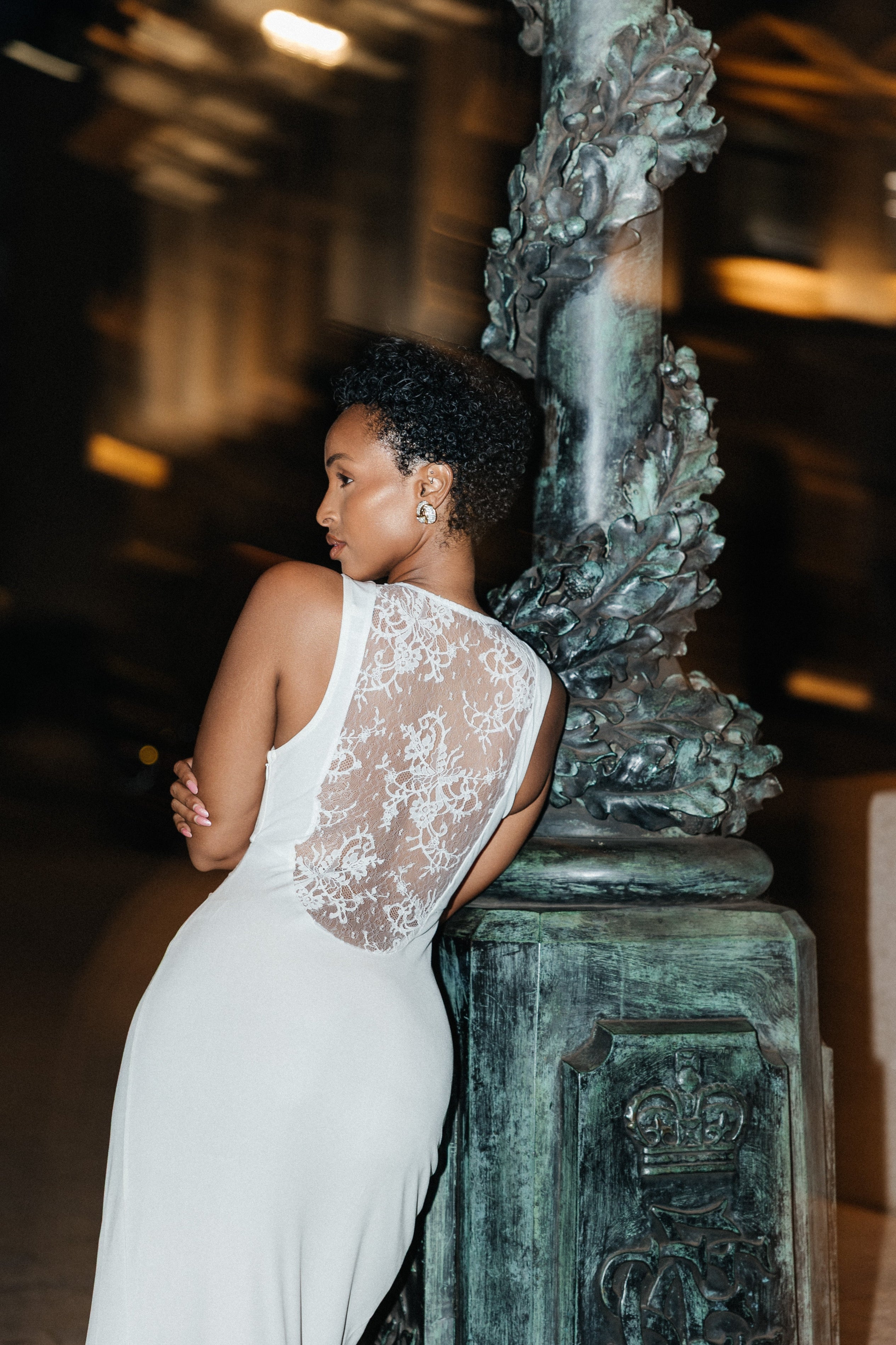 Woman in a white lace dress leaning against a decorative column at night.