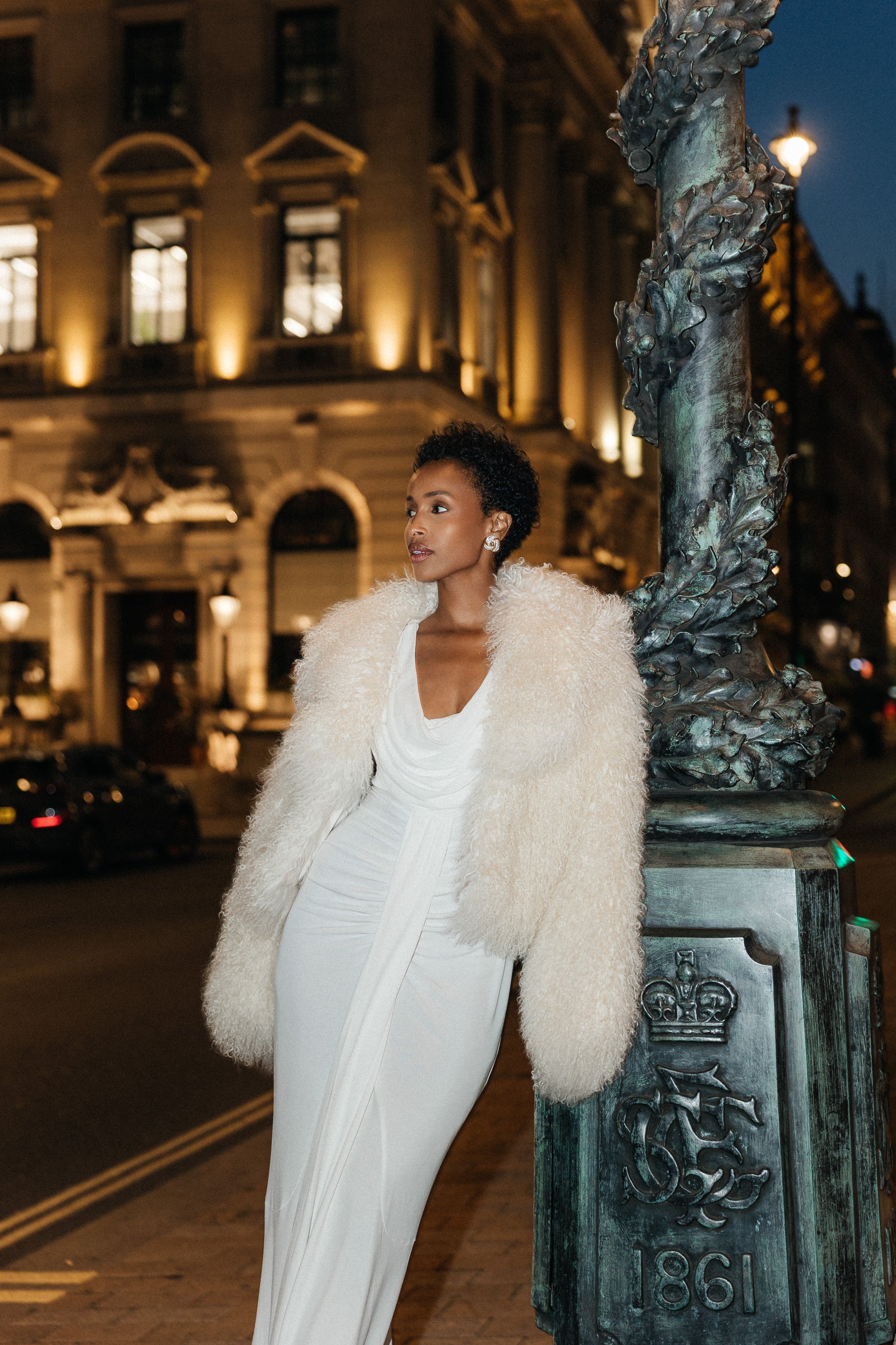 Woman in a white dress with a fur coat standing on a street at night.