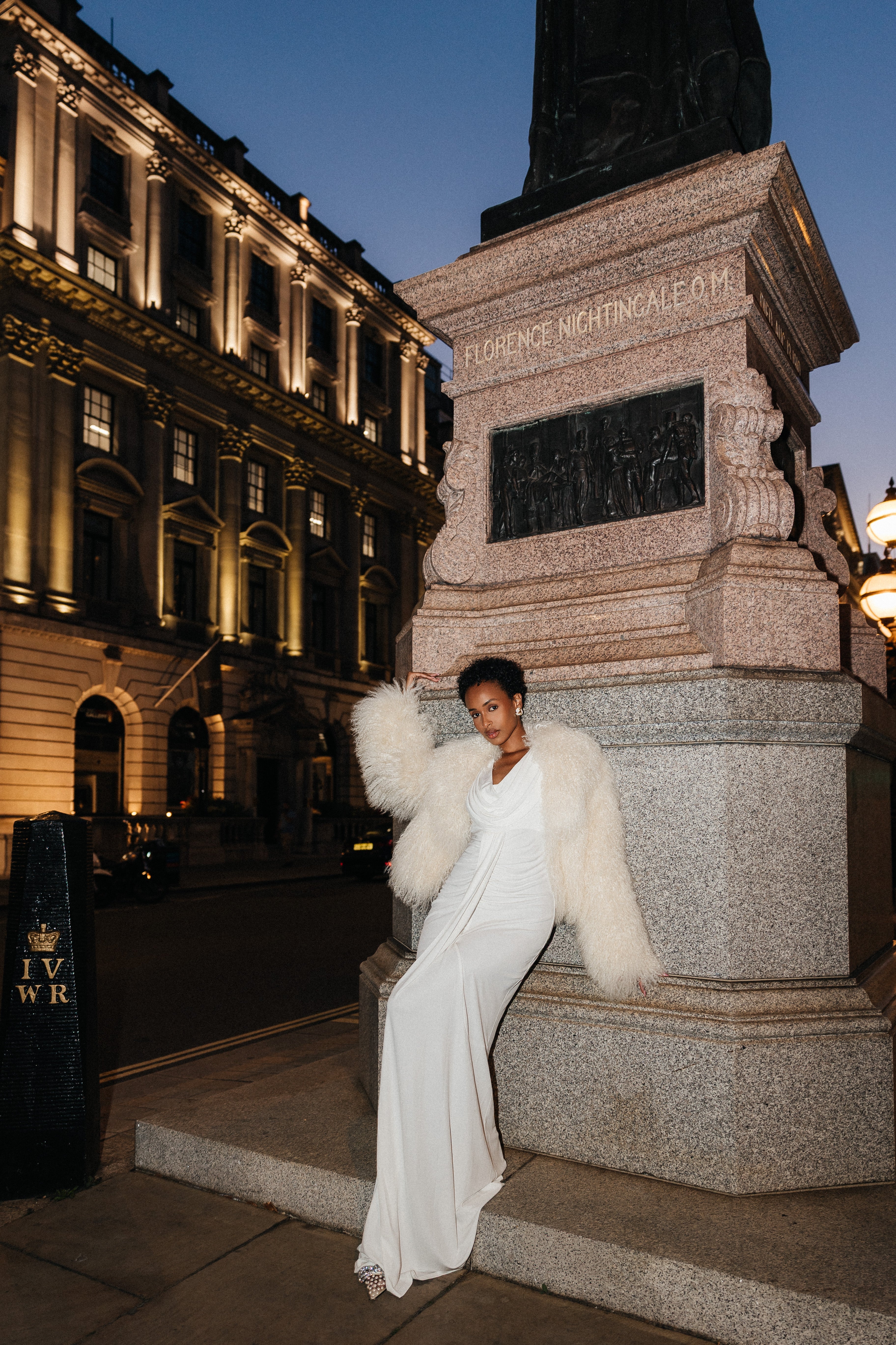 Person in a white outfit with fur accents standing in front of a stone monument at night.